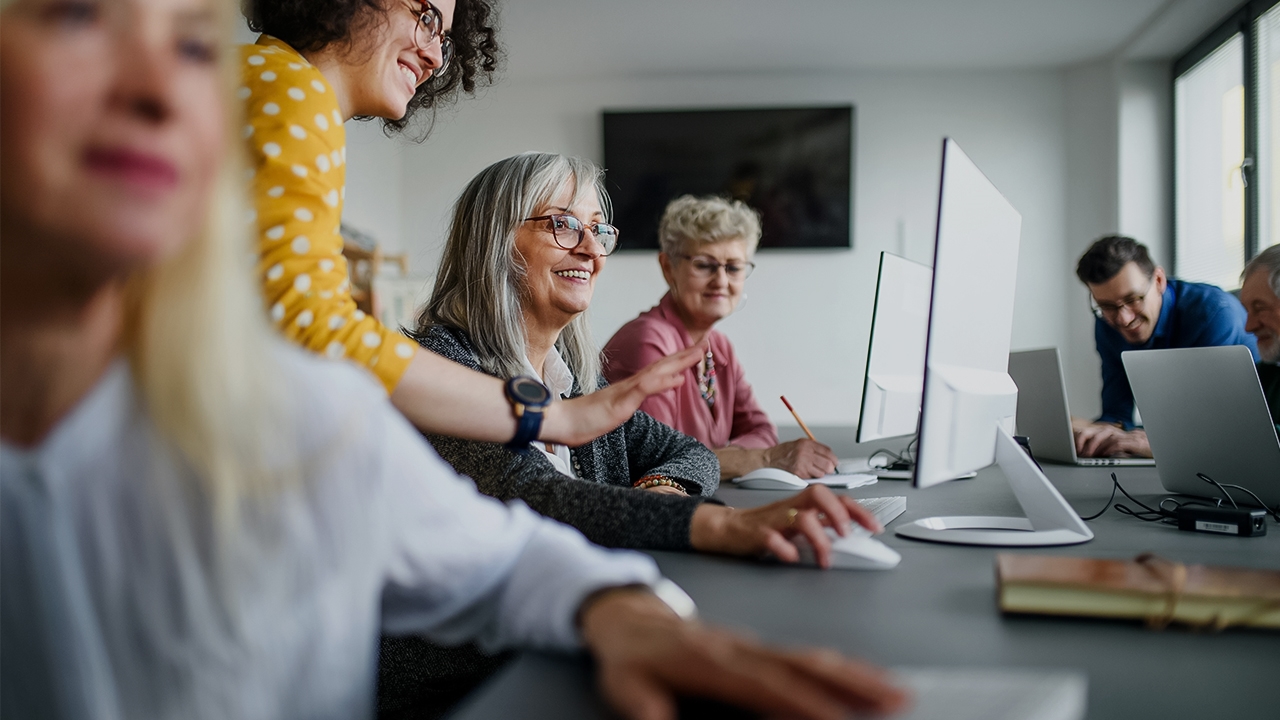 Mehrere Personen sitzen an einem Tisch vor einem Computer und Coaches stehen erklärend neben ihnen ©  Unsplash+ 