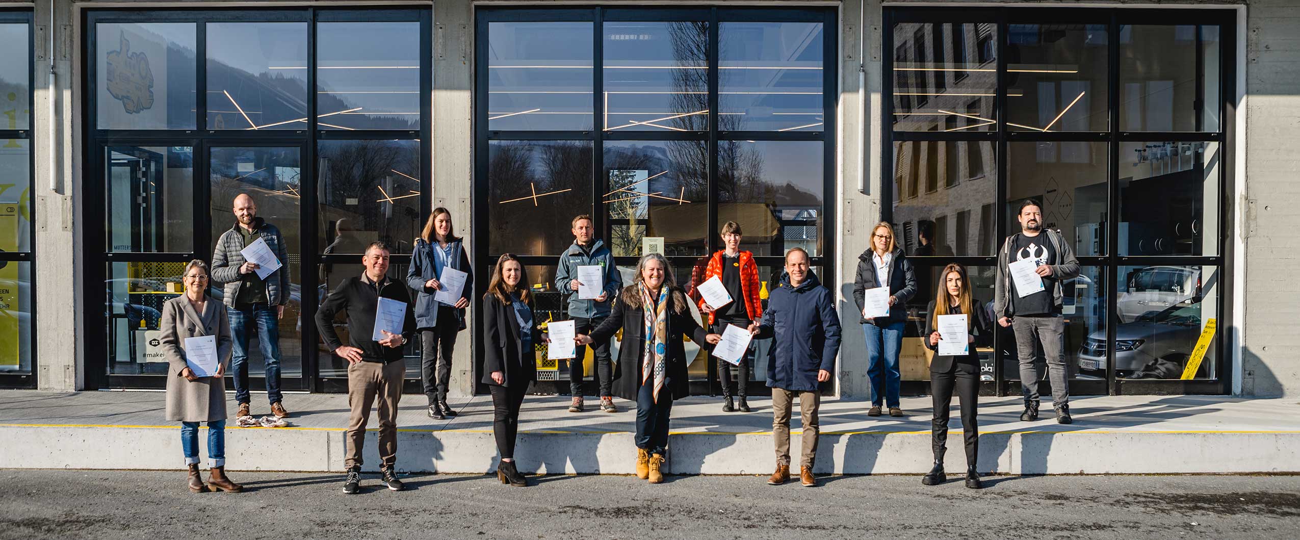 Eine Gruppe Menschen steht mit jeweils einem Ordner in der Hand vor einem großen, gläsernen Gebäude. © Mint4all Vorarlberg - Mint4all