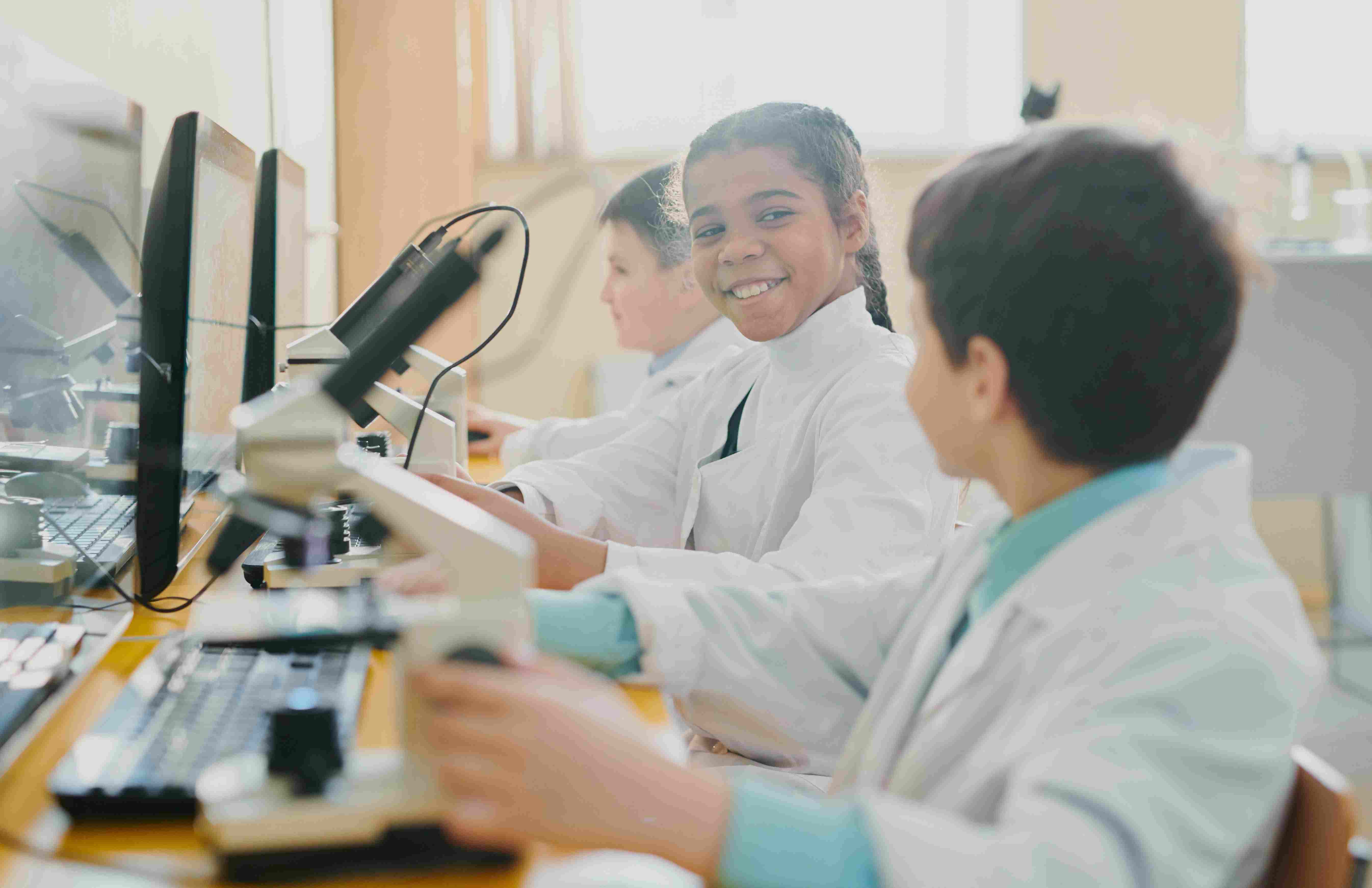 Drei Kinder, die in Laboruniform vor einem Computer und Mikroskop sitzen © Unsplash+